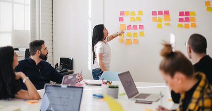 In einem gefüllten Meeting-Raum klebt eine Frau Post-Its an eine Wand. Andere Teilnehmende richten ihre Blicke auf sie.