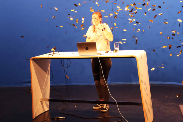 Moderatorin an einem Brückentisch in einem TV-Studio vor blauer Rückwand. Lametta in der Luft.