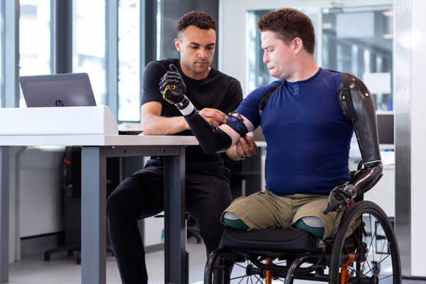 Ein Mann befestigt die Armprothese eines zweiten Mannes. Dieser sitzt im Rollstuhl.