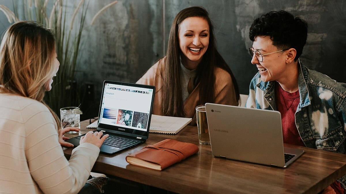 Lachende Schülerinnen sitzen an einem Tisch mit geöffneten Laptops.