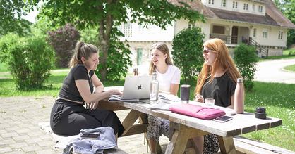 Drei Studentinnen arbeiten gemeinsam an einem Projekt. Sie sitzen im Freien an einer Holzbank und schauen gemeinsam in einen Laptop.