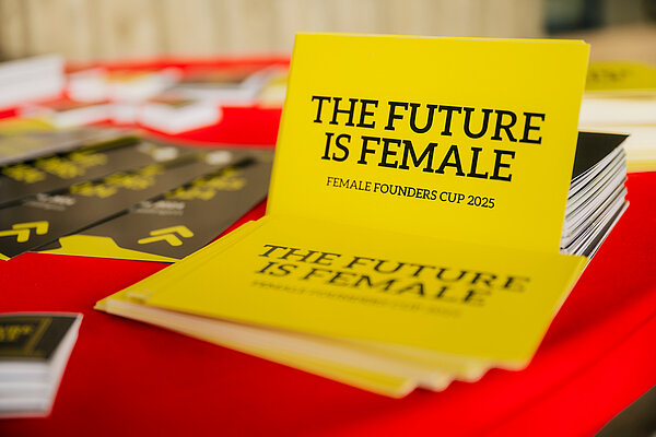 Key Visual zum Female Founders Cup 2025. Es liegen mehrere Postkarten auf einem Tisch mit rotem Untergrund. Text: THE FUTURE IS FEMALE, FEMALE FOUNDERS CUP 2025.