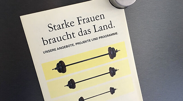 Flyer, der mit einem Magneten an einer grauen Wand befestigt ist. Aufschrift: "Starke Frauen braucht das Land."