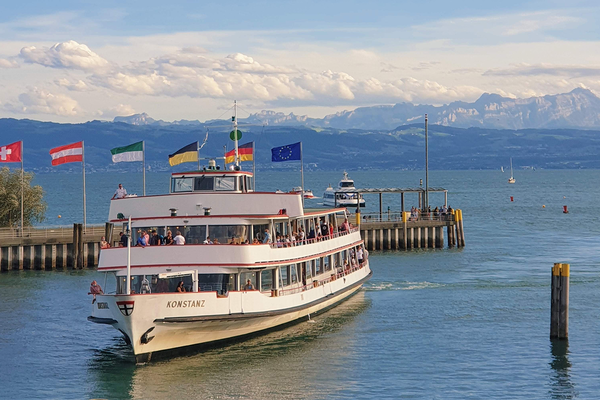 Ein Schiff fährt einen Hafen am Bodensee an. Im Hintergrund Landesflaggen von europäischen Ländern sowie Berge.
