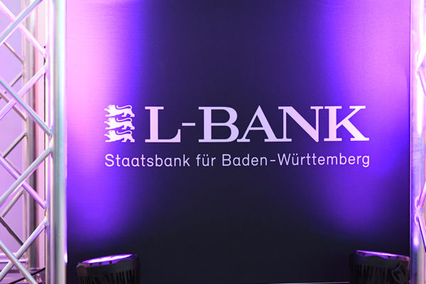 L-Bank-Logo in weiß auf schwarzem Textil, angestrahlt in magenta.