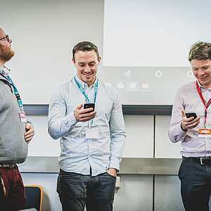 Drei Männer stehen nebeneinander und lachen während sie in ihr Handy schauen.