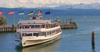 Ein Schiff fährt einen Hafen am Bodensee an. Im Hintergrund Landesflaggen von europäischen Ländern sowie Berge.