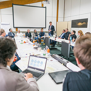 Mehrere Personen sitzen in einem Meetingraum. Eine Person steht und hält einen Vortrag.
