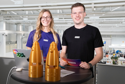 Eine Moderatorin und ein Moderator von NXTGN stehen an einem Stehtisch und lächeln in die Kamera; vor ihnen steht der Pokal für die Siegerinnen und Sieger der 14. Wettbewerbsrunde von ASAP.