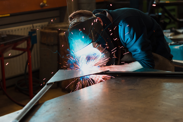 Ein Mann trägt einen Schweißhelm und übt in einer Werkstatt Schweißarbeiten an einem Metallteil aus. Zwischen dem Schweißgerät und dem Metallteil entstehen Funken. 