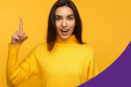 Frau im gelben Pullover steht vor einer gelben Rückwand und zeigt mit dem rechten Zeigefinger nach oben.