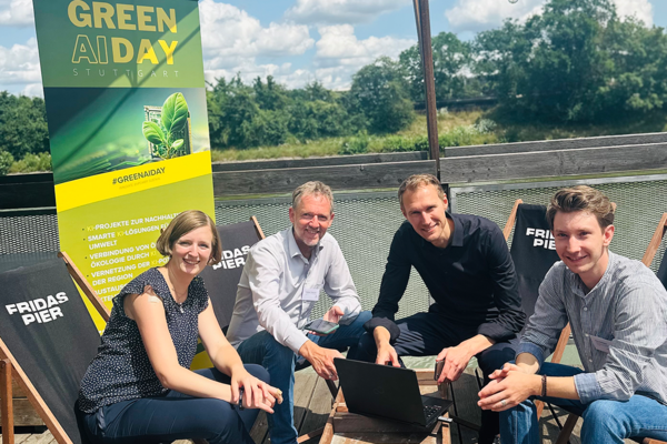 Vier Personen sitzen um einen Laptop herum auf dem Deck der Veranstaltungslocation Fridas Pier. Im Hintergrund ist ein Roll-up zur Veranstaltung Green AI Day Stuttgart zu sehen. Bildrechte: Landeshauptstadt Stuttgart.