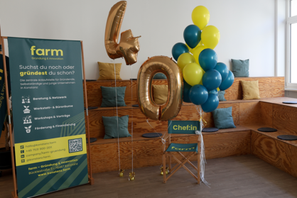 Innenaufnahme eines Veranstaltungsraums mit einer Sitztribüne. Auf den Sitzflächen liegen runde Sitzkissen und farbige Dekokissen (gelb und grün). Im Vordergrund befinden sich goldene Luftballons in Form der Zahl „40“ sowie ein Strauß gelber und grüner Luftballons. Links steht ein Roll-up mit Informationen über „farm – Gründung & Innovation“. Auf einem Klappstuhl steht ein Schild mit der Aufschrift „Chef:in“. Bildrechte: farm.