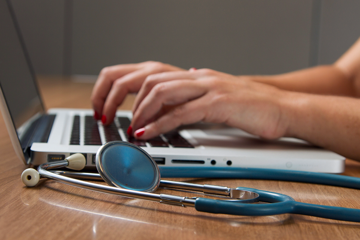 Aufgeklappter Laptop, an dem eine Person mit rot lackierten Fingernägeln arbeitet. Neben der Tastatur befindet sich ein Stethoskop.