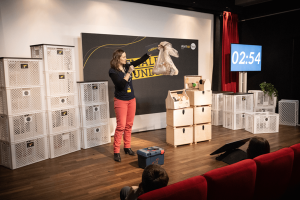 Eine Gründerin präsentiert ihre Geschäftsidee auf der Bühne beim Female Founders Cup 2022. Sie hält einen gelben Sack mit Plastikmüll in den Händen.