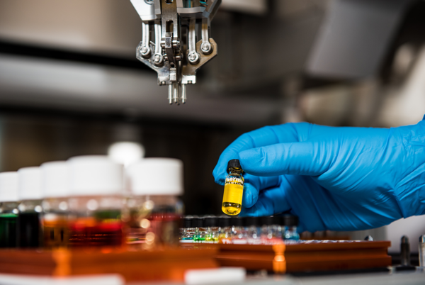 Hand mit blauem Gummihandschuh hält ein Glasgefäß mit einer Laborprobe.