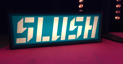 Mit Licht hinterleuchtetes Logo des Start-up Festivals SLUSH - Weiße Buchstaben auf petrolfarbenem Hintergrund.